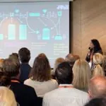 Presentatör håller föredrag om bostadsmarknad inför publik i konferenssal, diagram visas på projektor