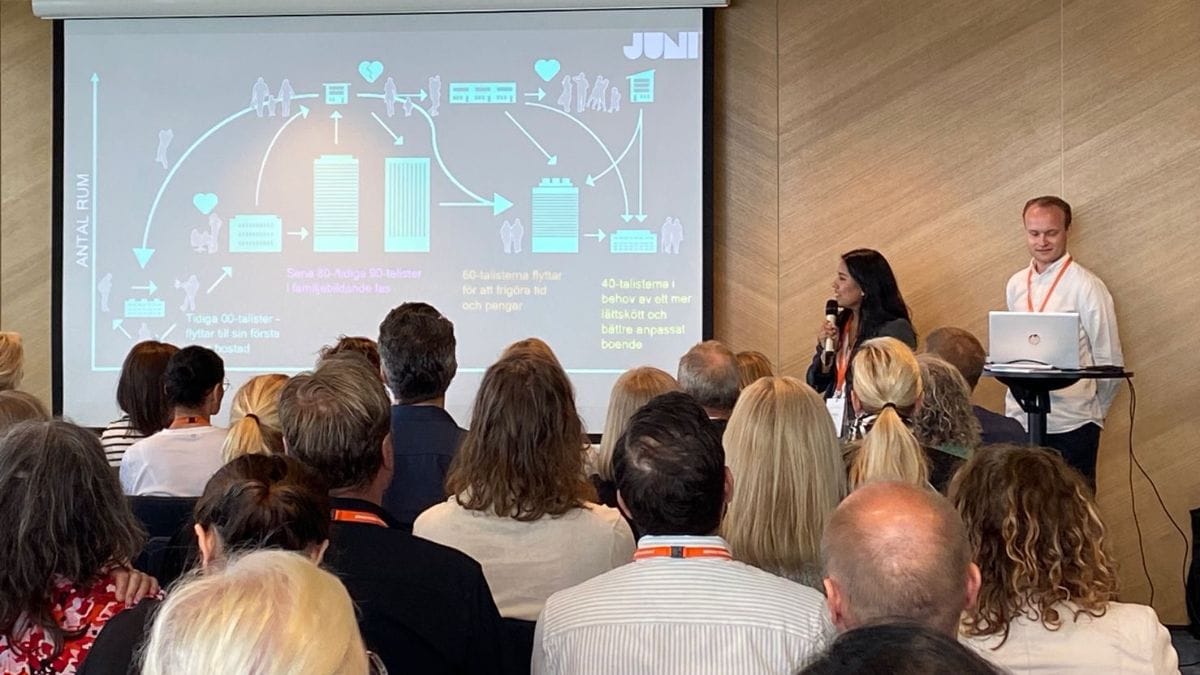 Presentatör håller föredrag om bostadsmarknad inför publik i konferenssal, diagram visas på projektor