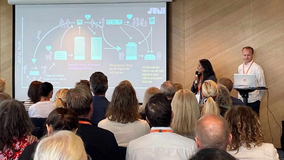 Presentatör håller föredrag om bostadsmarknad inför publik i konferenssal, diagram visas på projektor