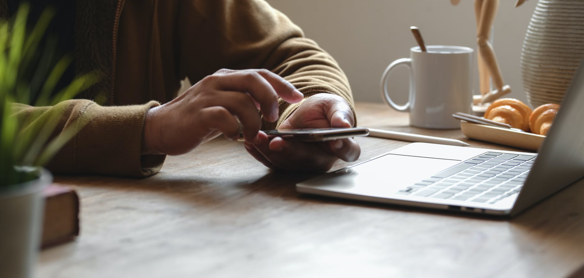 Person arbetar med smartphone vid laptop på kontorsskrivbord med kaffe och frallor
