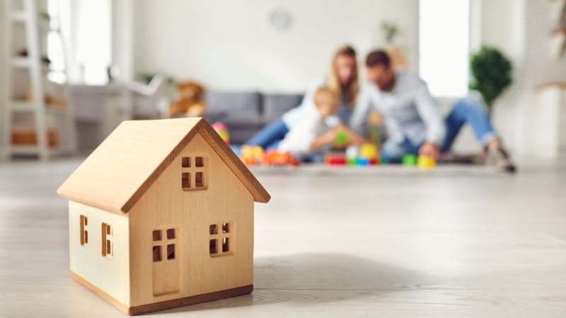 Trähusmodell i förgrunden med familj som leker på golvet i bakgrunden, symboliserar bostadsdrömmar och familjeliv