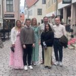Sex personer står tillsammans på kullerstensgata i historisk stadsmiljö med affärer och restauranger