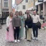 Sex personer står tillsammans på kullerstensgata i historisk stadsmiljö med affärer och restauranger